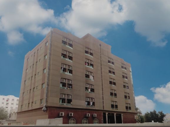 Exterior view of a multi-story beige building with air conditioners, set against a partly cloudy sky.